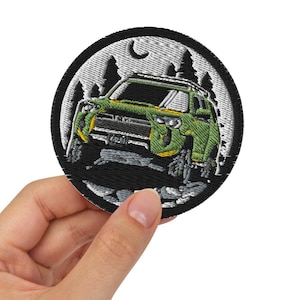 Pode incluir: Um remendo bordado com um carro verde dirigindo por uma floresta à noite, com uma lua crescente no céu.