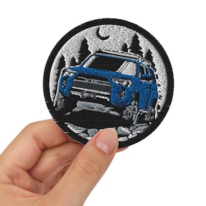 Könnte beinhalten: Ein blauer gestickter Patch mit einem weißen Mond, Bäumen und einem blauen SUV, der auf einem felsigen Weg fährt. Der Patch ist rund und hat einen schwarzen Rand.