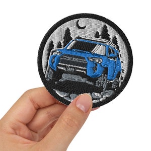 May include: A black and white embroidered patch featuring a blue car driving through a forest scene. The patch includes a crescent moon and stars in the sky.