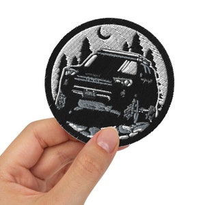 May include: A black and white embroidered patch depicting a car driving through a forest at night. The patch features a crescent moon and stars in the background.