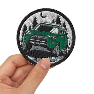 Könnte beinhalten: Ein grüner gestickter Aufnäher mit schwarzem Hintergrund, der einen grünen Geländewagen zeigt, der nachts durch einen Wald fährt. Der Aufnäher hat einen weißen Mond und weiße Bäume.