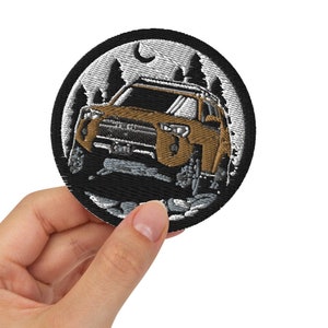 May include: A black and white embroidered patch featuring a brown car driving through a forest at night. The patch includes a moon and stars in the sky.