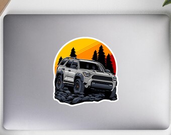 Puede incluir: Un SUV gris conduce por un terreno rocoso con una puesta de sol de fondo. El SUV tiene una parrilla negra y ruedas negras. La puesta de sol es naranja y amarilla.