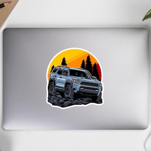 Pode incluir: Um SUV cinza dirigindo por um terreno rochoso com um pôr do sol ao fundo. O SUV tem uma grade preta e rodas pretas. O pôr do sol é laranja e amarelo com uma silhueta de árvores na frente.