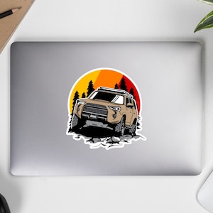 Könnte beinhalten: Ein brauner SUV mit einem Dachgepäckträger fährt über ein felsiges Gelände mit einem Sonnenuntergang im Hintergrund. Die Sonne ist ein Farbverlauf von Orange, Gelb und Rot. Im Hintergrund befinden sich Bäume.