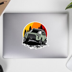 May include: A green SUV with a roof rack drives through a rocky terrain with a sunset in the background. The sunset is a gradient of orange, red, and yellow. There are trees in the background.