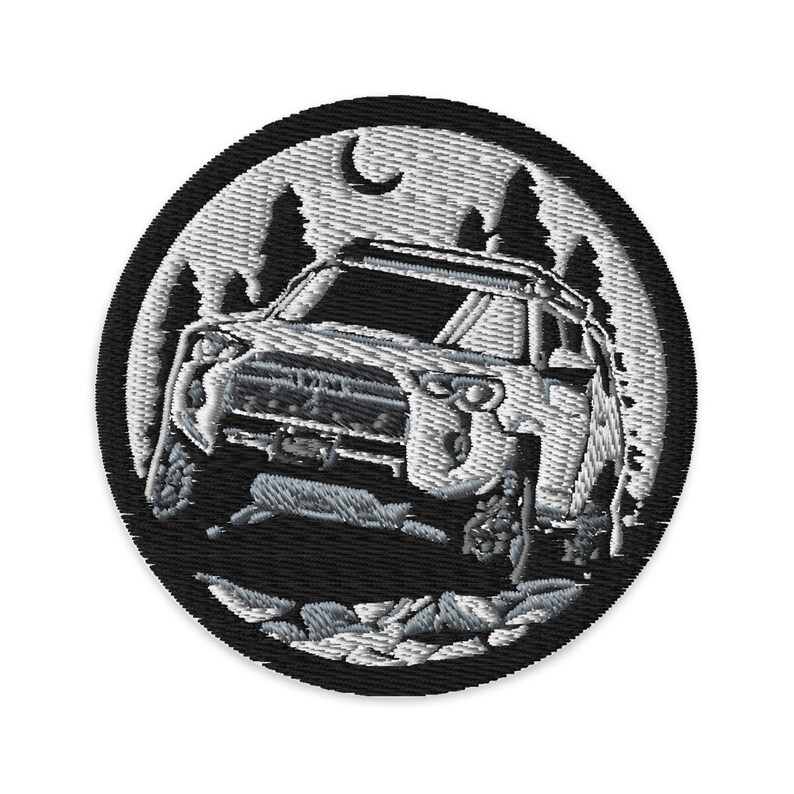 Pode incluir: Um remendo bordado branco com fundo preto que representa um SUV branco dirigindo por uma floresta &agrave; noite. O SUV tem um porta-malas no teto e uma lua &eacute; vis&iacute;vel no c&eacute;u.