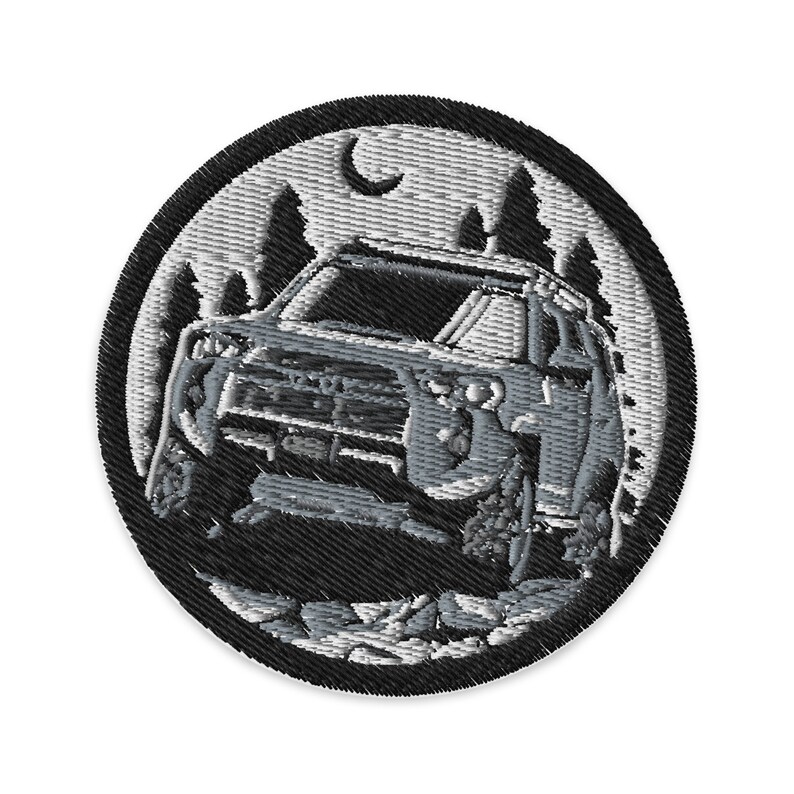 Puede incluir: Un parche bordado en blanco y negro de un SUV plateado conduciendo por un bosque de noche. El parche es circular y tiene un borde negro.