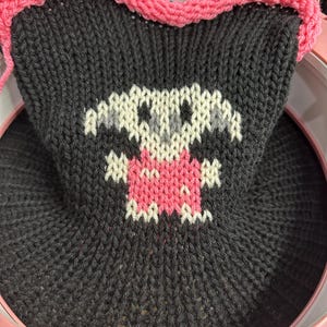 May include: A black knitted fabric with a pink border, featuring a white and gray knitted design of a creature with a pink heart on its chest.