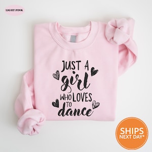 May include: A light pink sweatshirt with the text "Just a girl who loves to dance" in black lettering with hearts around the text.