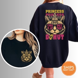 May include: A navy blue sweatshirt featuring a cat wearing sunglasses and a crown. The text on the back reads "PRINCESS DONUT" with "THIS IS AN OUTRAGE!" on the cat's face. A smaller version of the design is on the front.
