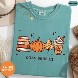May include: A seafoam green t-shirt featuring a graphic print of a stack of books, a pumpkin, a maple leaf, and a coffee cup with whipped cream. The text "cozy season" is printed below the graphic, accompanied by small star icons.