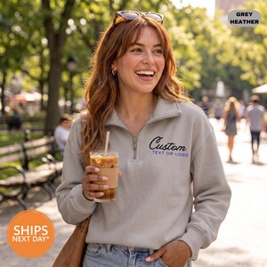 May include: A grey heather quarter-zip sweatshirt featuring a collar, long sleeves, and the text "Custom TEXT OR LOGO" embroidered in black and blue on the left chest. The garment is worn with blue denim jeans and sunglasses.