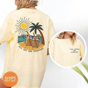 May include: A light yellow t-shirt with a graphic on the back featuring a sun, palm tree, and two iced coffee drinks. The text "ALL DAY AND EVERYDAY" is below the graphic. The front of the shirt has the text "ICED COFFEE AND SUNSHINE".