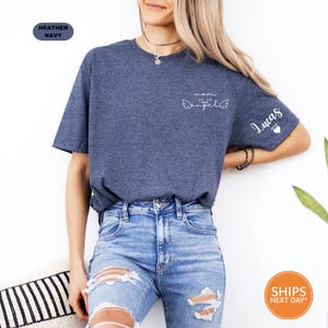 A woman wearing a blue t-shirt with a graphic design and jeans stands against a white background.