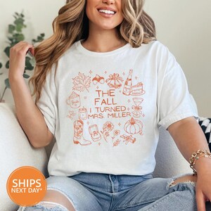 May include: A white short-sleeved t-shirt featuring orange line art graphics including a maple leaf, acorn, bells, champagne, cake, mug, candle, cowboy boot, ring box, flowers, pumpkin, and cinnamon sticks with the text THE FALL I TURNED MRS. MILLER.