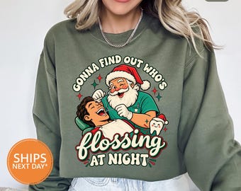 Könnte beinhalten: Ein militärgrüner Rundhals-Pullover mit einer Grafik, die den Weihnachtsmann als Zahnarzt bei der Untersuchung eines Patienten zeigt. Der Text lautet "GONNA FIND OUT WHO'S FLOSSING AT NIGHT" mit einem kleinen Zahn mit Weihnachtsmütze.