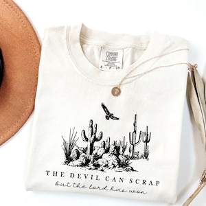 May include: A cream colored t-shirt with a black graphic of a desert scene with cacti and a flying bird. The text "THE DEVIL CAN SCRAP but the lord has won" is printed below the graphic.
