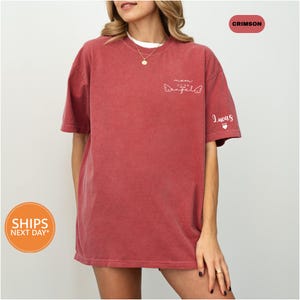 A woman wearing a red t-shirt with a graphic design on it, standing against a white background.