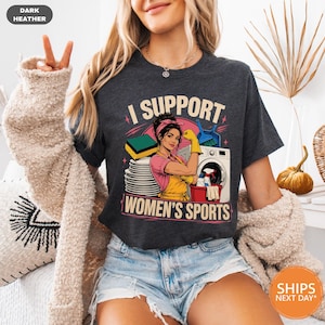 May include: A dark heather gray short-sleeve t-shirt featuring a graphic print of a person wearing a yellow apron and gloves, surrounded by cleaning supplies like a sponge, dishes, and a washing machine, with the text I SUPPORT WOMEN'S SPORTS.