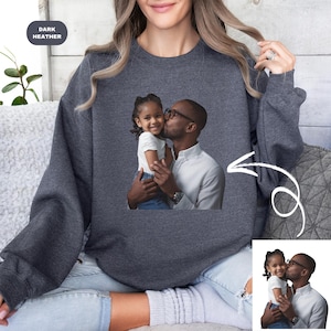May include: A dark heather grey sweatshirt with a photo of a father and daughter. The father is kissing the daughter on the cheek. The photo is printed on the front of the sweatshirt.
