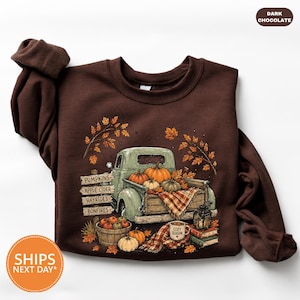May include: A dark chocolate brown long-sleeve sweatshirt featuring a graphic of a vintage light green pickup truck loaded with pumpkins. A wooden sign lists PUMPKINS, APPLE CIDER, HAYRIDES, and BONFIRES. A mug labeled COZY SEASON sits nearby.