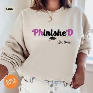 May include: A light beige sweatshirt with the text "PhinisheD" in black and pink lettering. Underneath the text is a black graduation cap with a pink tassel and the text "Dr. Jonas" in black.