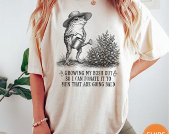 May include: An ivory t-shirt featuring a black graphic of a frog wearing a hat and holding a watering can next to a bush. The text reads: GROWING MY BUSH OUT SO I CAN DONATE IT TO MEN THAT ARE GOING BALD. The shirt has short sleeves and a crew neckline.