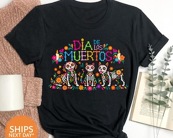 May include: A black t-shirt featuring a colourful graphic of a dog, a cat, and another dog designed as sugar skulls. The text "DIA DE LOS MUERTOS" is printed in bright, multi-coloured letters above the animals, surrounded by floral accents.