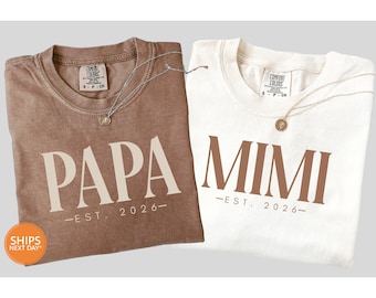 May include: Two t-shirts, one brown and one white, with the words "PAPA" and "MIMI" printed in large letters. Below each word is "-EST. 2026-". Each shirt has a small tag and a necklace.