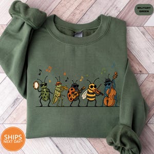 May include: A military green crewneck sweatshirt featuring a graphic print of five insects playing musical instruments. The insects include a ladybug with a tambourine, a grasshopper with a trumpet, a ladybug with a guitar, a bee with a violin, and a beetle with a double bass, all wearing sunglasses.