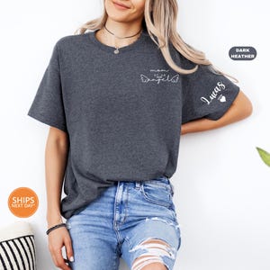A woman wearing a gray t-shirt with a cat design and jeans stands against a white wall, smiling.