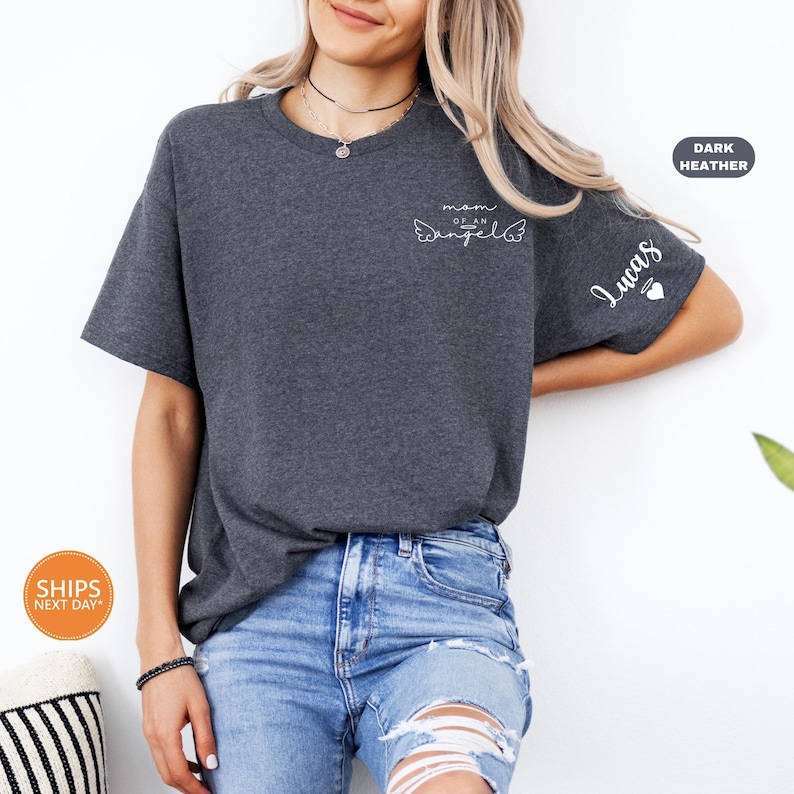 A woman wearing a gray t-shirt with a cat design and jeans stands against a white wall, smiling.