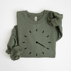 May include: A green sweatshirt with a black clock graphic on the front. The clock has a single hand pointing to the right.