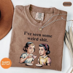 May include: Espresso-colored t-shirt with the text "I've seen some weird shit." features an illustration of two women at a table. The shirt has a Comfort Colors tag and a silver necklace. A brown hat is visible in the background.