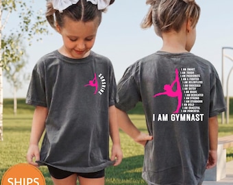 May include: A dark heather grey t-shirt featuring a pink gymnast silhouette on the front left chest with the word SUNSHINE. The back displays a large pink gymnast silhouette with the text I AM SMART, TOUGH, PASSIONATE, FIGHTER, RELENTLESS, OBSESSED, GUTSY, BRAVE, DEDICATED, STRONG, STUBBORN, BOLD, GRACEFUL, POWERFUL, I AM GYMNAST.