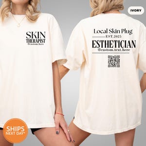 May include: Ivory-colored t-shirt with black text. The front reads "SKIN THERAPIST" and "@custom.here". The back says "Local Skin Plug EST.2025 ESTHETICIAN @custom.text.here" with a QR code. The shirt is a casual style.