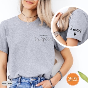 May include: Sport grey t-shirt with the text "mom of an angel" and angel wings graphic. The sleeve has the name "Lucas" and a heart. The shirt is made of a soft material.