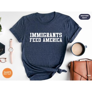 May include: Heather navy blue t-shirt with the text "IMMIGRANTS FEED AMERICA" in white block letters. The shirt is displayed on a white surface with a watch, eyeglasses, a coffee cup, and a brown leather bag.