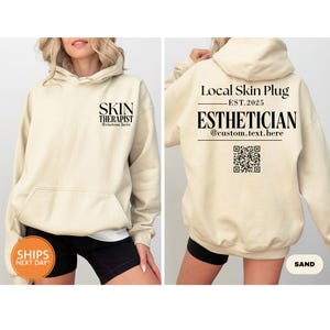 May include: Sand-colored hoodie with the text "SKIN THERAPIST" on the front and "Local Skin Plug EST.2025 ESTHETICIAN" on the back. The hoodie has a drawstring hood and a front pocket. The text is in black.