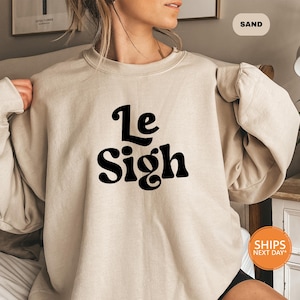 May include: A beige sweatshirt with the text "Le Sigh" printed in black on the front.