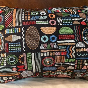 Puede incluir: Una almohada negra con un patrón geométrico colorido. El patrón incluye círculos, cuadrados, triángulos y líneas en tonos de rojo, azul, verde, amarillo y marrón.