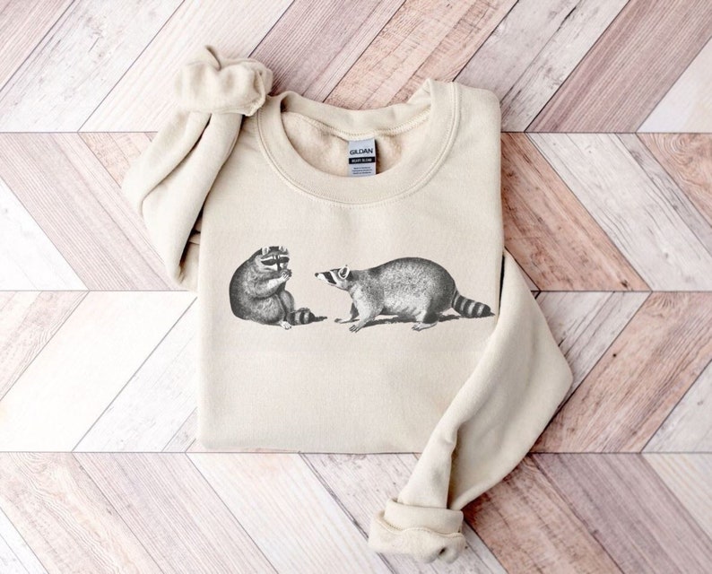May include: A beige sweatshirt with a black and white illustration of two raccoons on the front. The raccoons are facing each other and one is holding a piece of food.