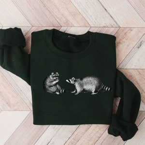 May include: A dark green sweatshirt with a graphic print of two raccoons. The raccoons are facing each other, one is sitting and the other is standing.