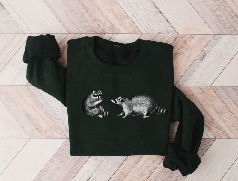 May include: A dark green sweatshirt with a graphic print of two raccoons. The raccoons are facing each other, one is sitting and the other is standing.