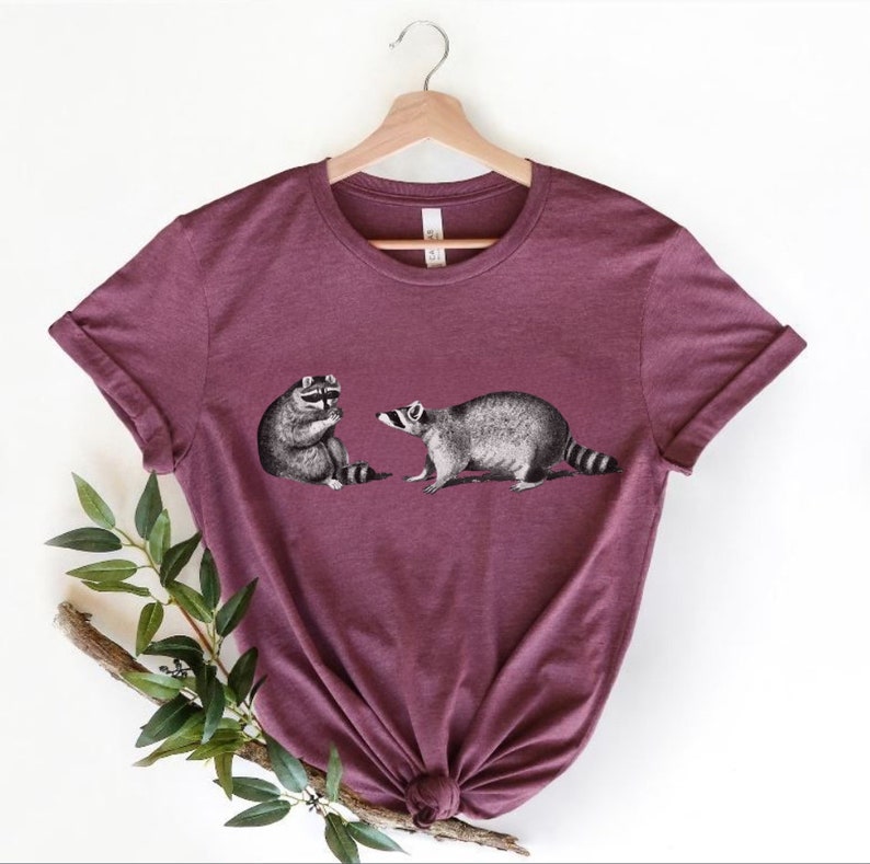 May include: A maroon t-shirt with a graphic of two raccoons. The raccoon on the left is sitting down and the raccoon on the right is standing up.
