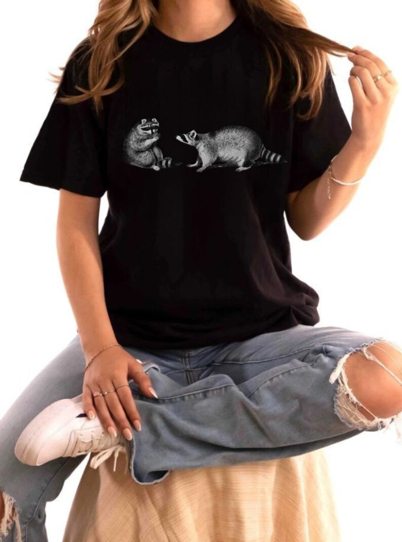 May include: A black t-shirt with a graphic print of two raccoons facing each other. The raccoons are drawn in a vintage style.