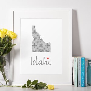 Puede incluir: Una impresión enmarcada en blanco con un contorno gris del estado de Idaho con un diseño estampado en gris y blanco. La palabra "Idaho" está escrita en cursiva gris con un pequeño corazón rojo al lado.