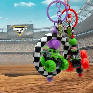 May include: A Monster Jam keyring with two miniature monster trucks, one green and one black, with red and green wheels. The keyring includes a chequered ring, purple accents, and heart-shaped charms. The background shows a stadium with the Monster Jam logo.