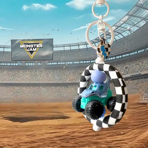 May include: A Monster Jam keyring featuring a blue monster truck toy with black wheels, a checkered arch, and a heart-shaped charm. The keyring has a white ring and a blue and white lightning bolt charm. The background shows a stadium and the Monster Jam logo.
